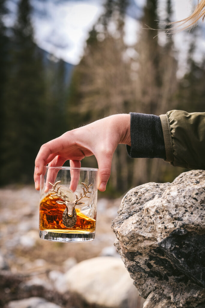 Swirling a glass of whiskey with a deer head on in in the mountains.